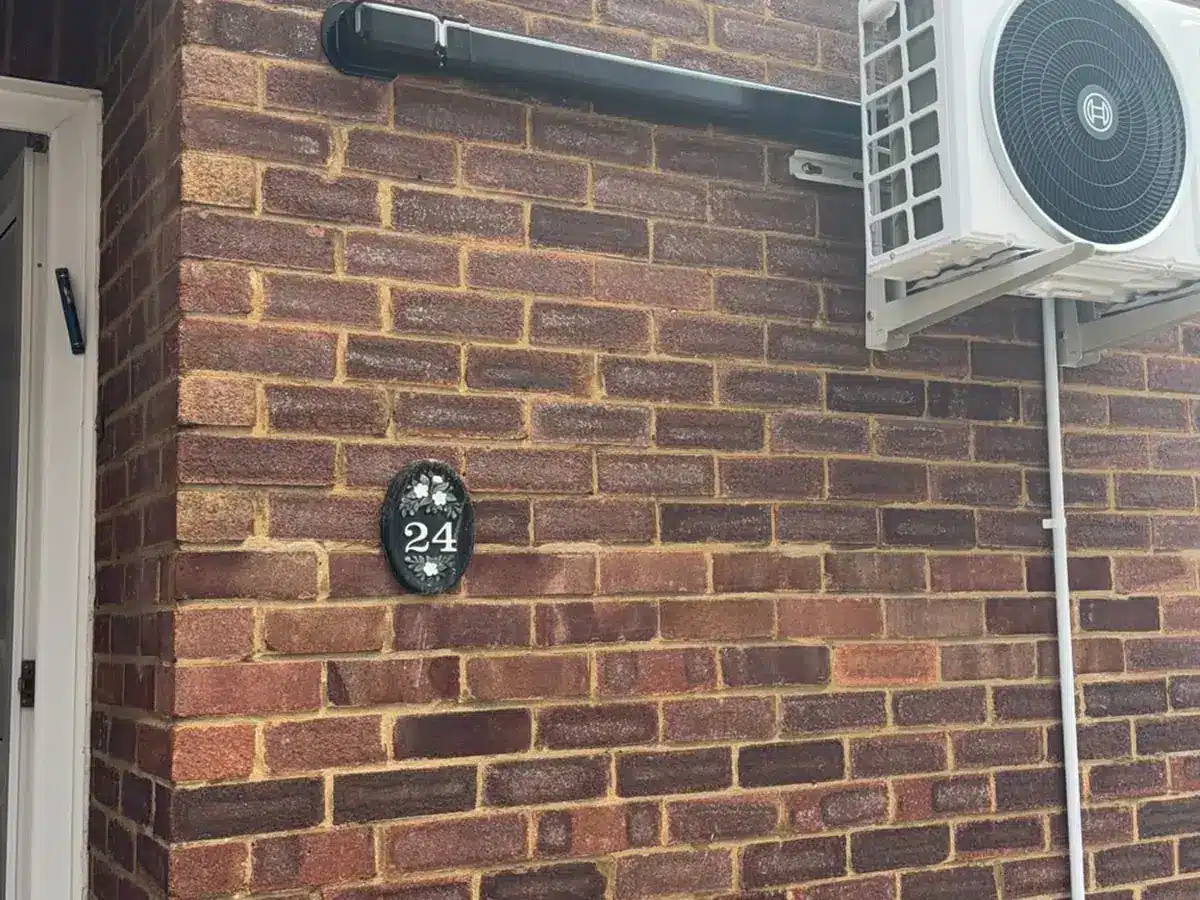 Outdoor unit on wall brackets with pipe trunking along a brick wall above a house number plaque