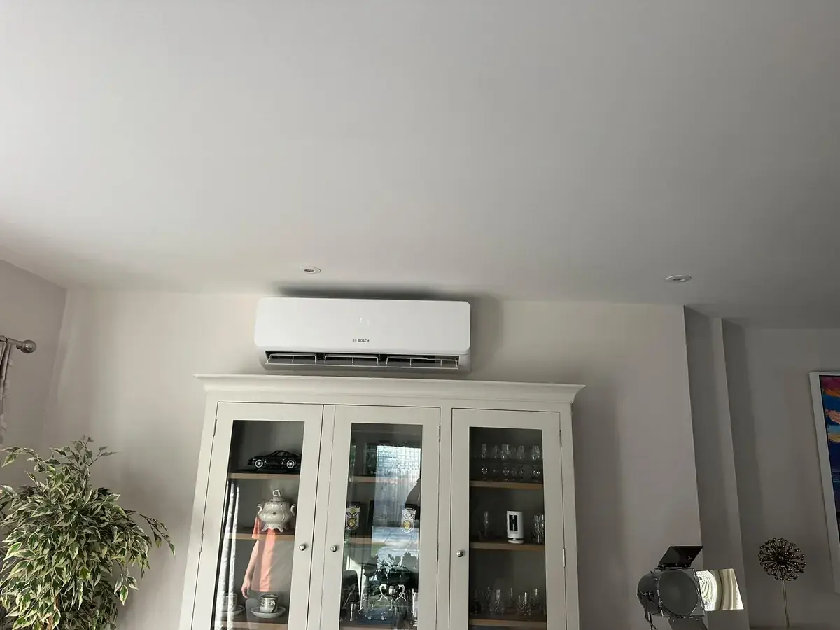 White wall-mounted air conditioning unit above a glass-fronted dresser in a living room