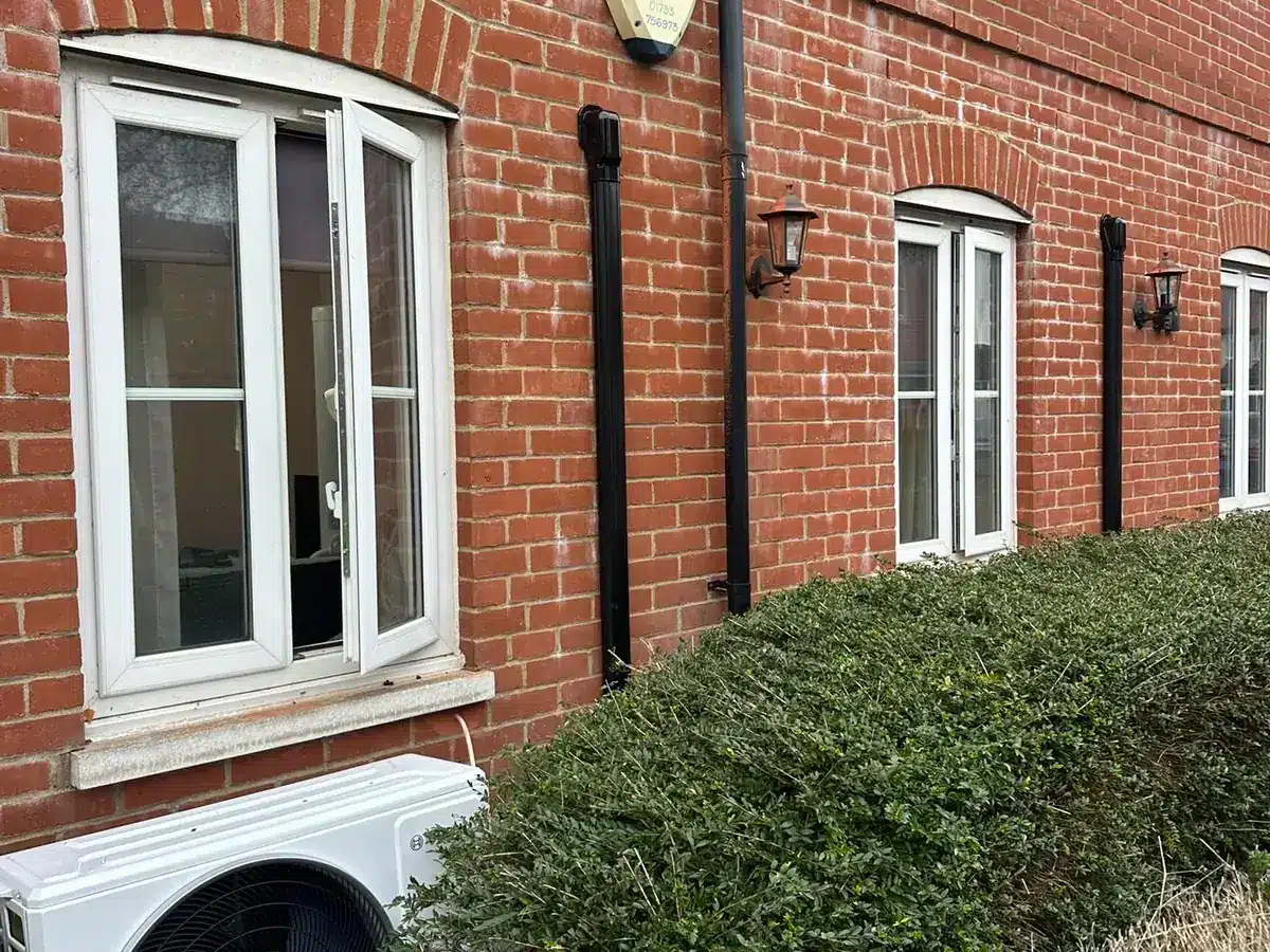 Outdoor unit at the base of a red-brick building behind a hedge