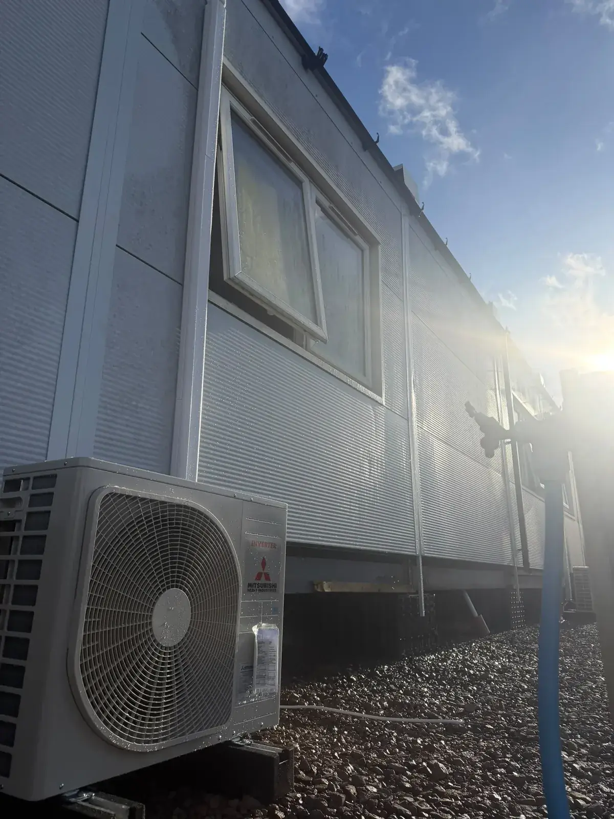 Mitsubishi Electric outdoor unit on gravel beside a clad building in bright sunlight