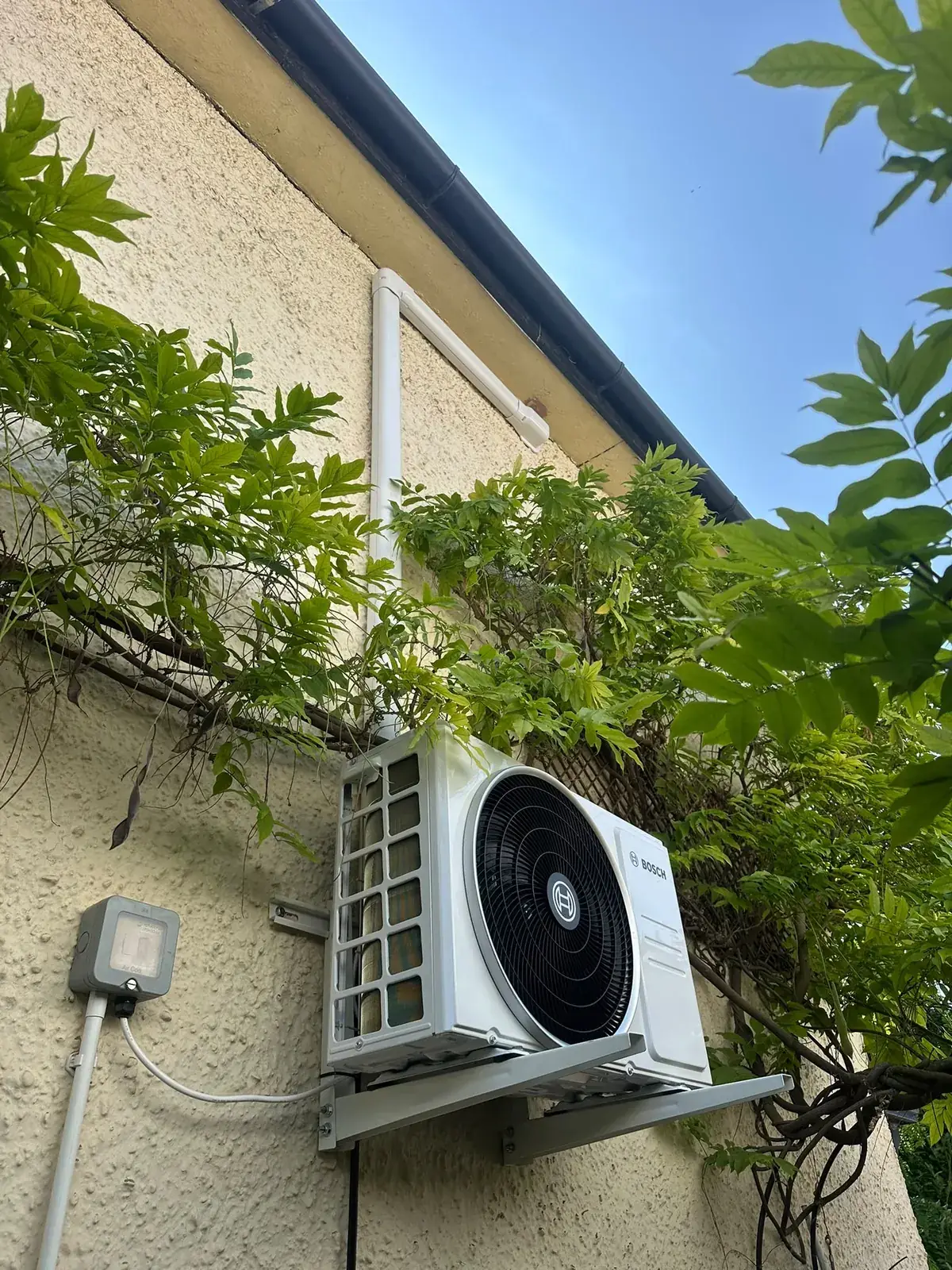 Bosch outdoor unit on a wall bracket among climbing plants on a rendered house wall